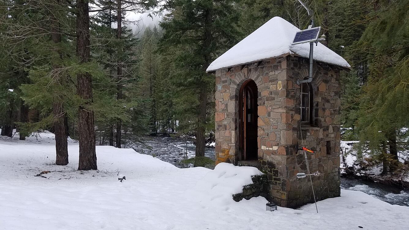 A small stone building sits on a snowy river bank