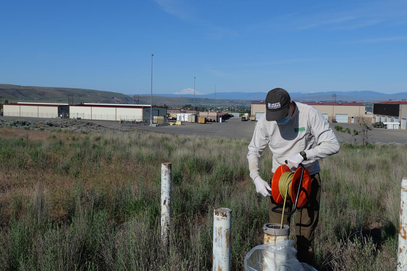 Scientist lowers tape into well in a field with warehouse
