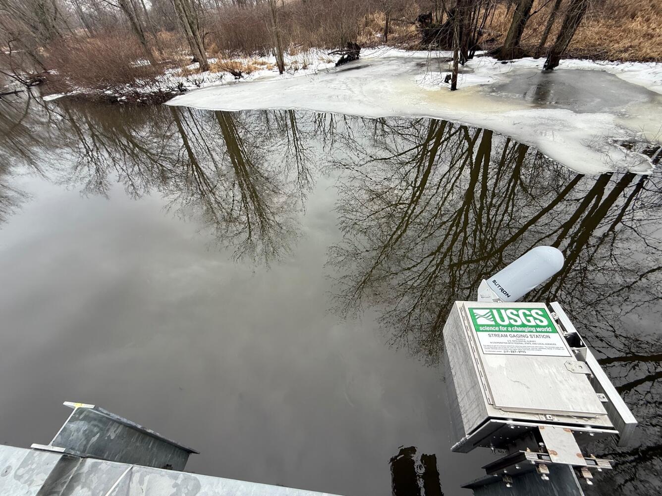 A streamgage housing is mounted on the side of a bridge over a creek. Ice covers the shoreline.