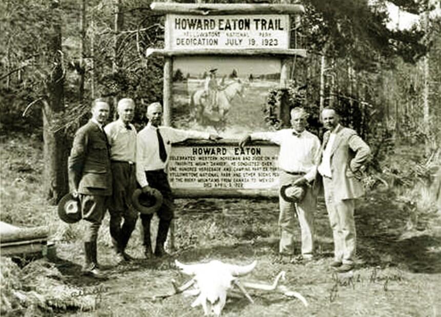 Five men in 1920s-era suits stand in front of a wooden sign for Howard Eaton Trail; forest behind and bison skull in front
