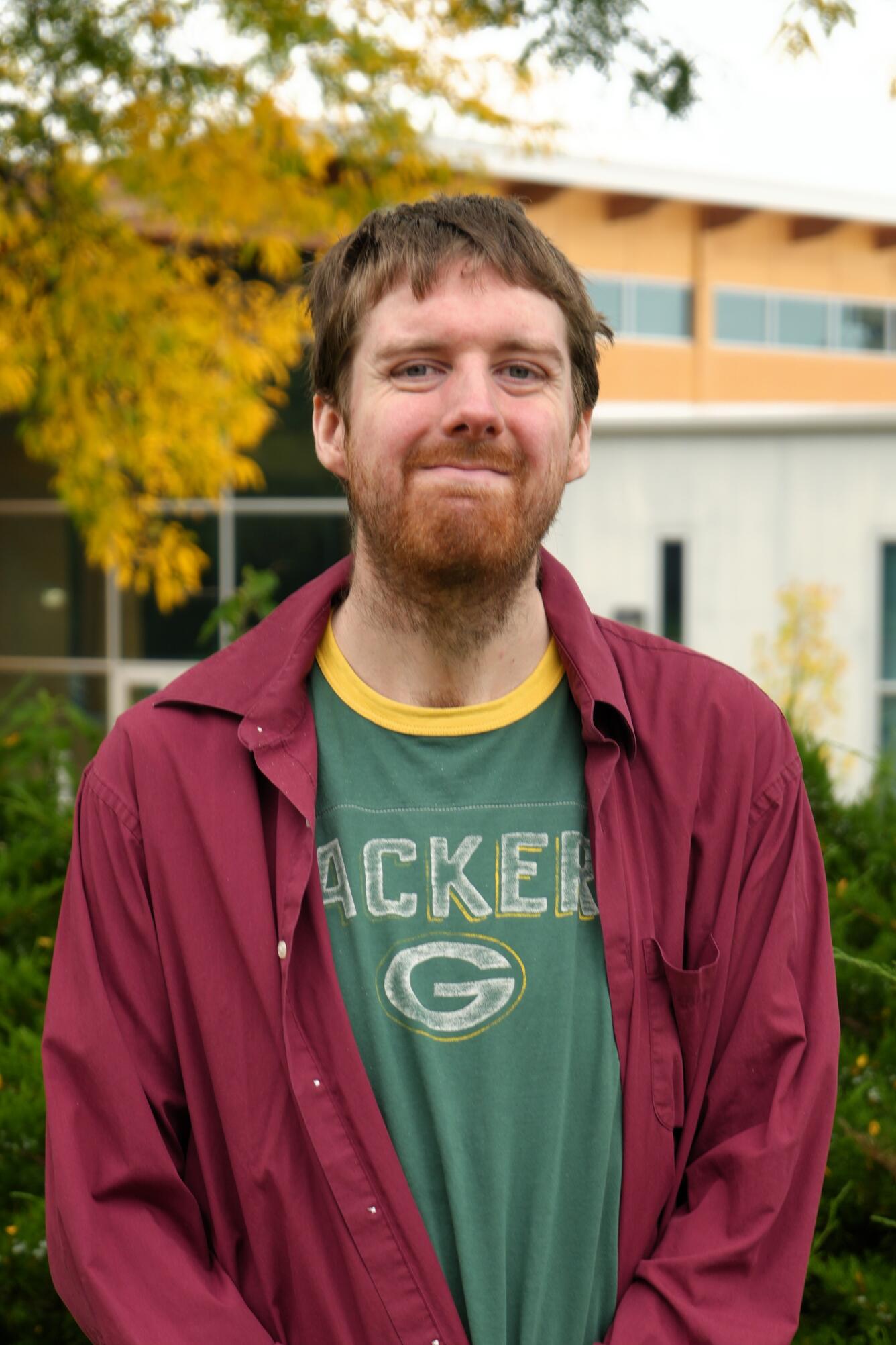Man smiling standing in front of building and tree with yellow leaves.