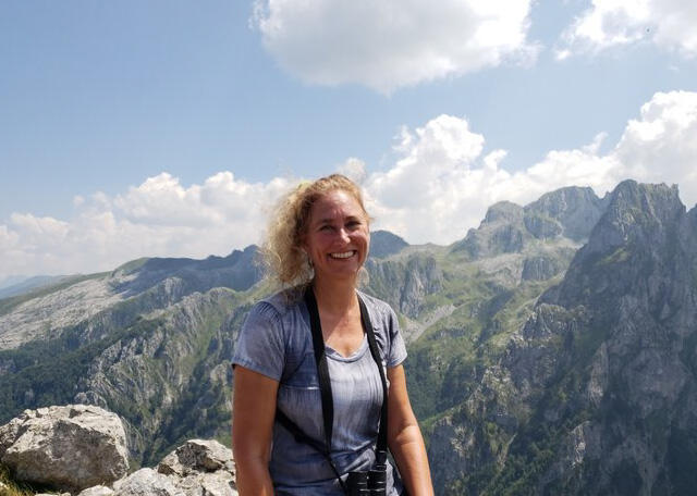 A woman with blonde hair smiles towards the camera with mountains behind her