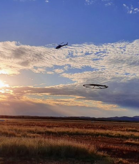 Helicopter seen aloft, towing a round hoop used to image geology.