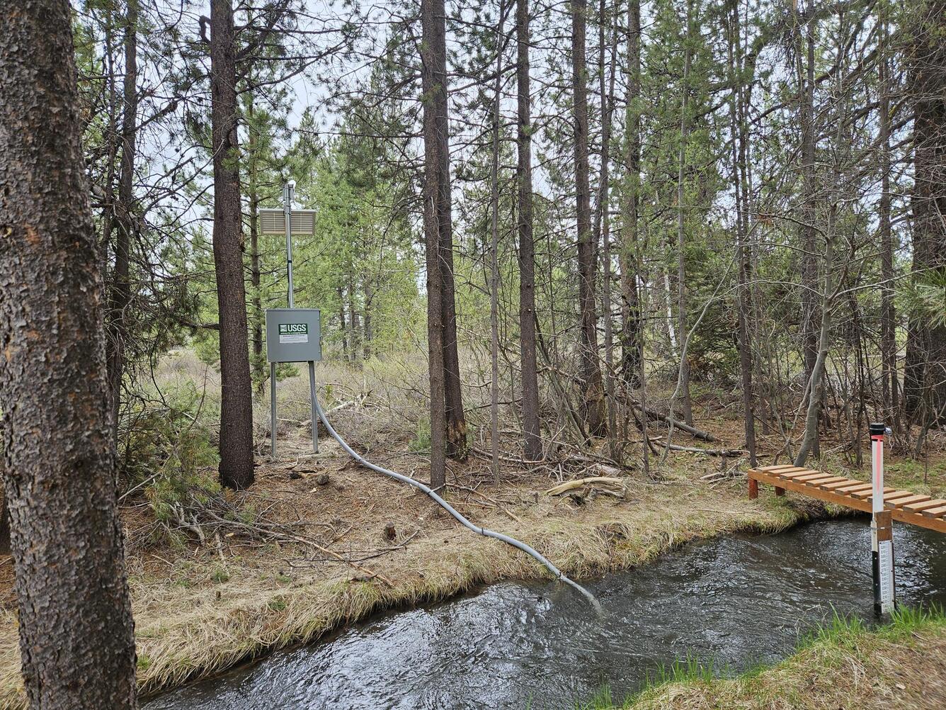 narrow stream with yellow grass, tree lined banks. Grey metal box houses monitoring equipment, solar panel and antenna.