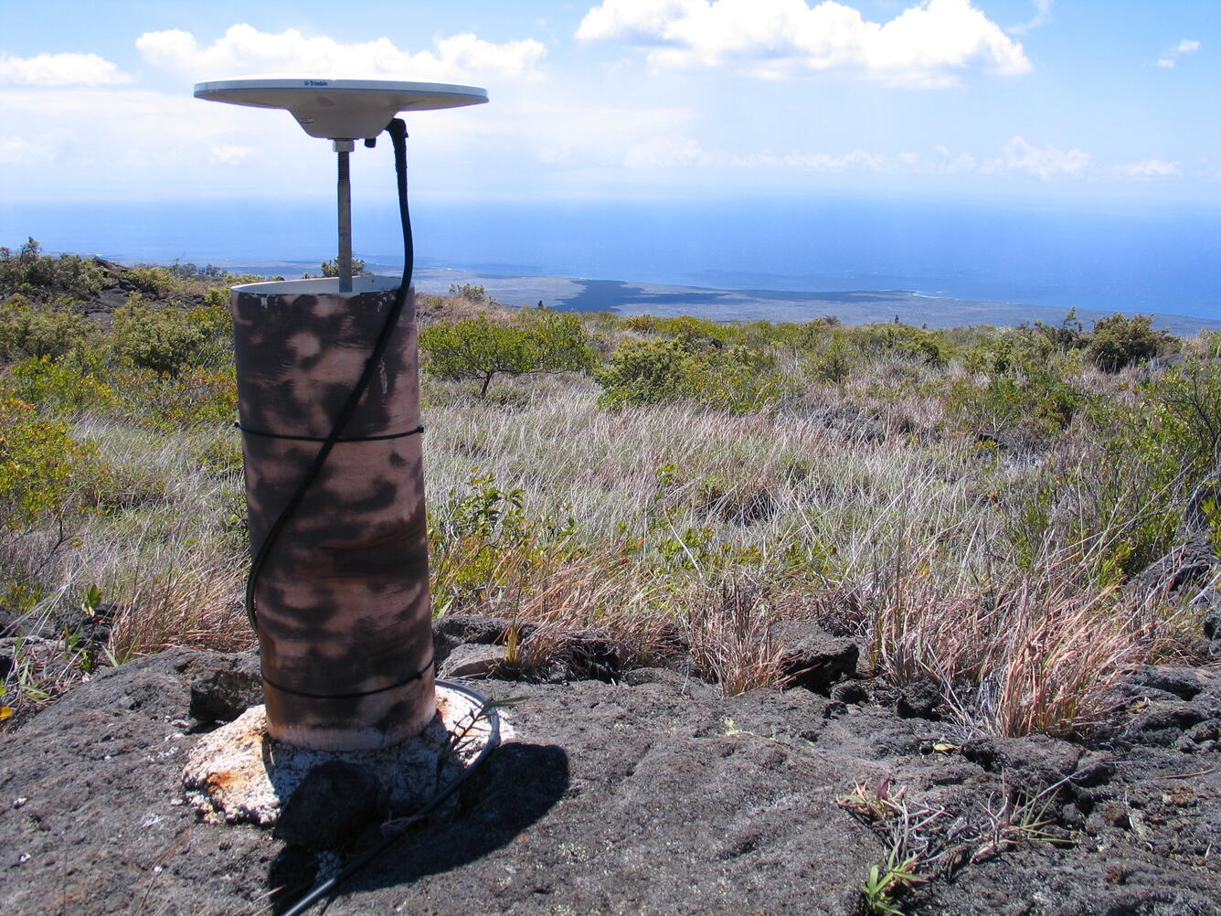Color photograph of volcano monitoring device with grass in the foreground and ocean in the background