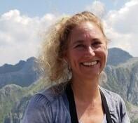 A person with blonde hair smiles towards the camera with mountains and blue sky in the background