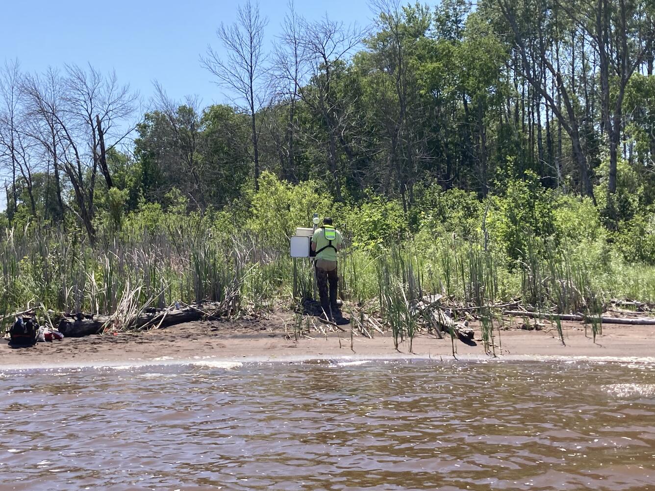 Hydrologic technician stands on shoreline and installs equipment in a streamgage housing