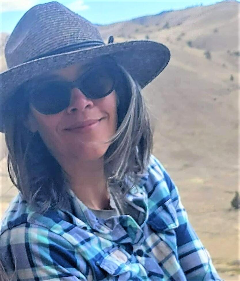 Amanda Garcia in sun glasses and sun hat poses against a backdrop of rolling yellow hills in John Day Oregon