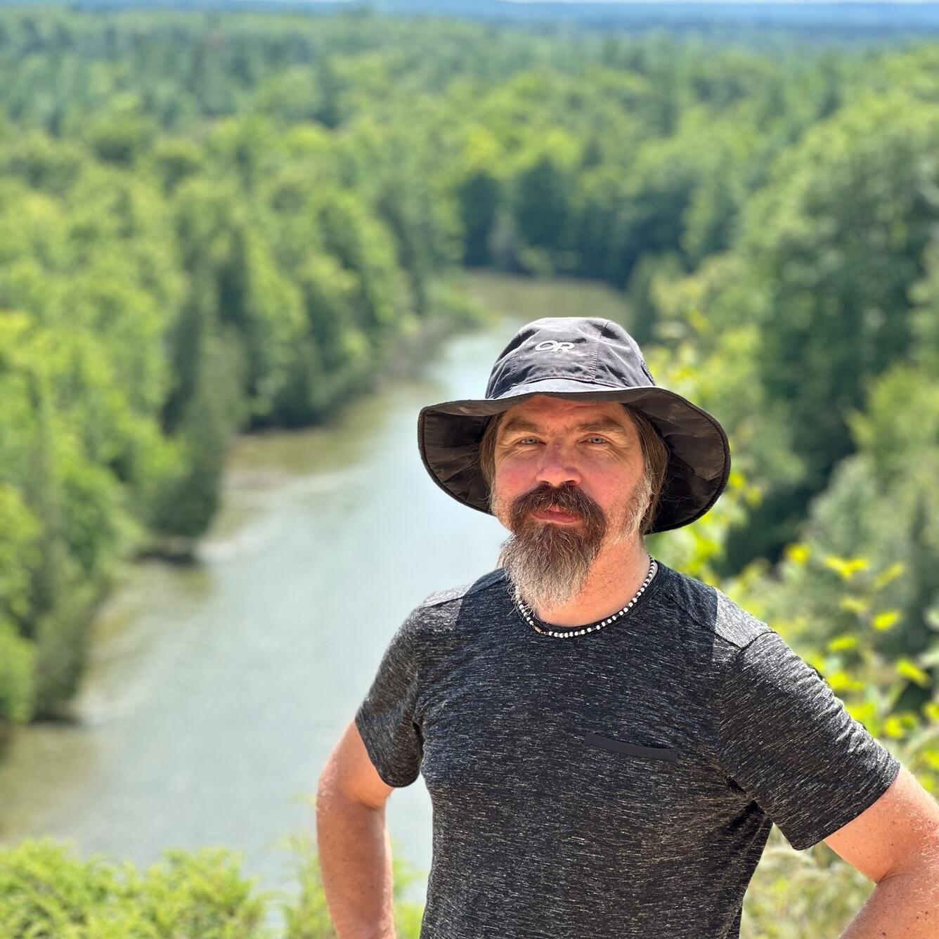 A bearded man in a hat poses in front of a river surrounded by trees.