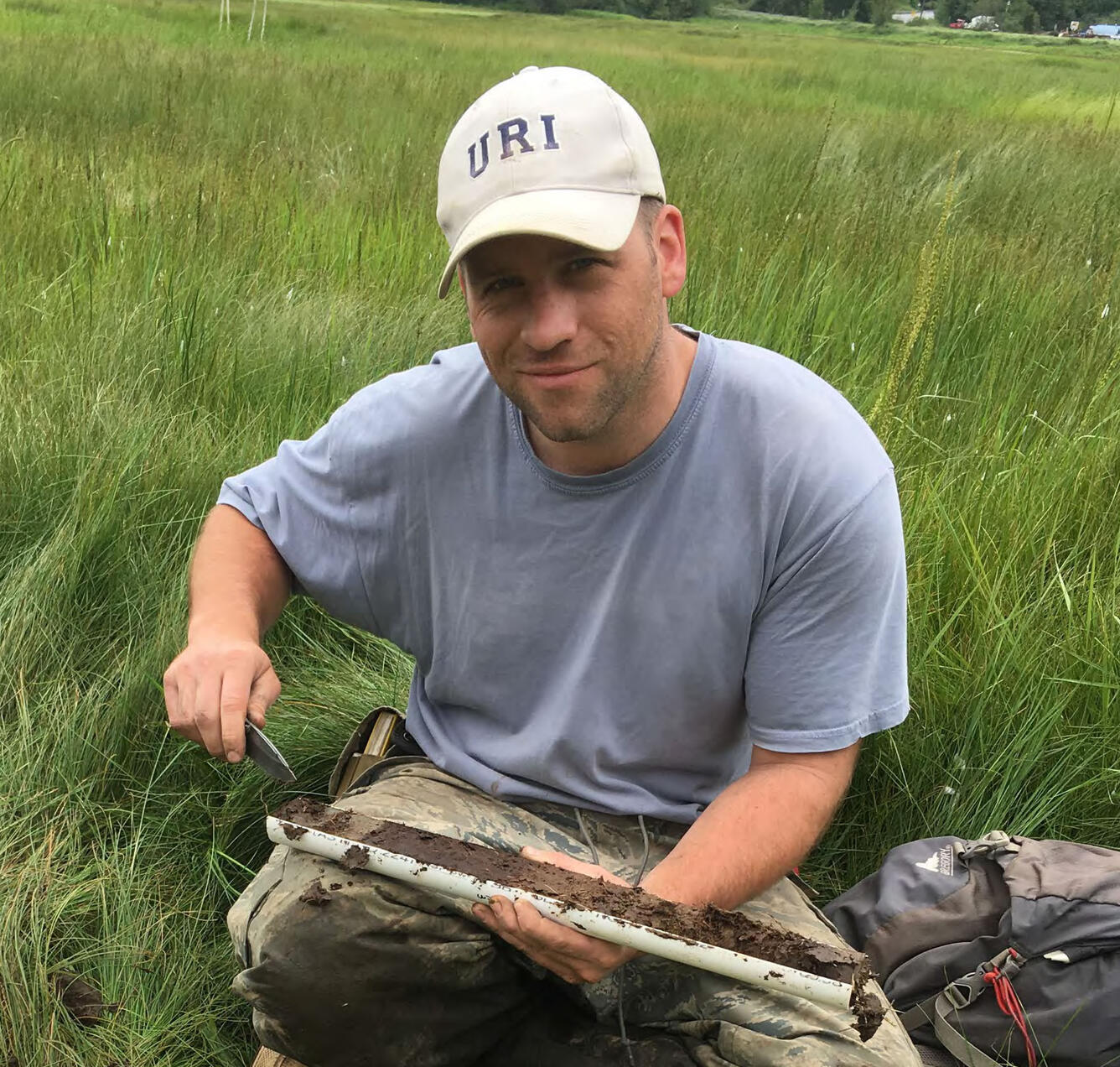 A smiling man, kneeling in a grassy field, holds a plastic tube cut open to show the sediment inside.