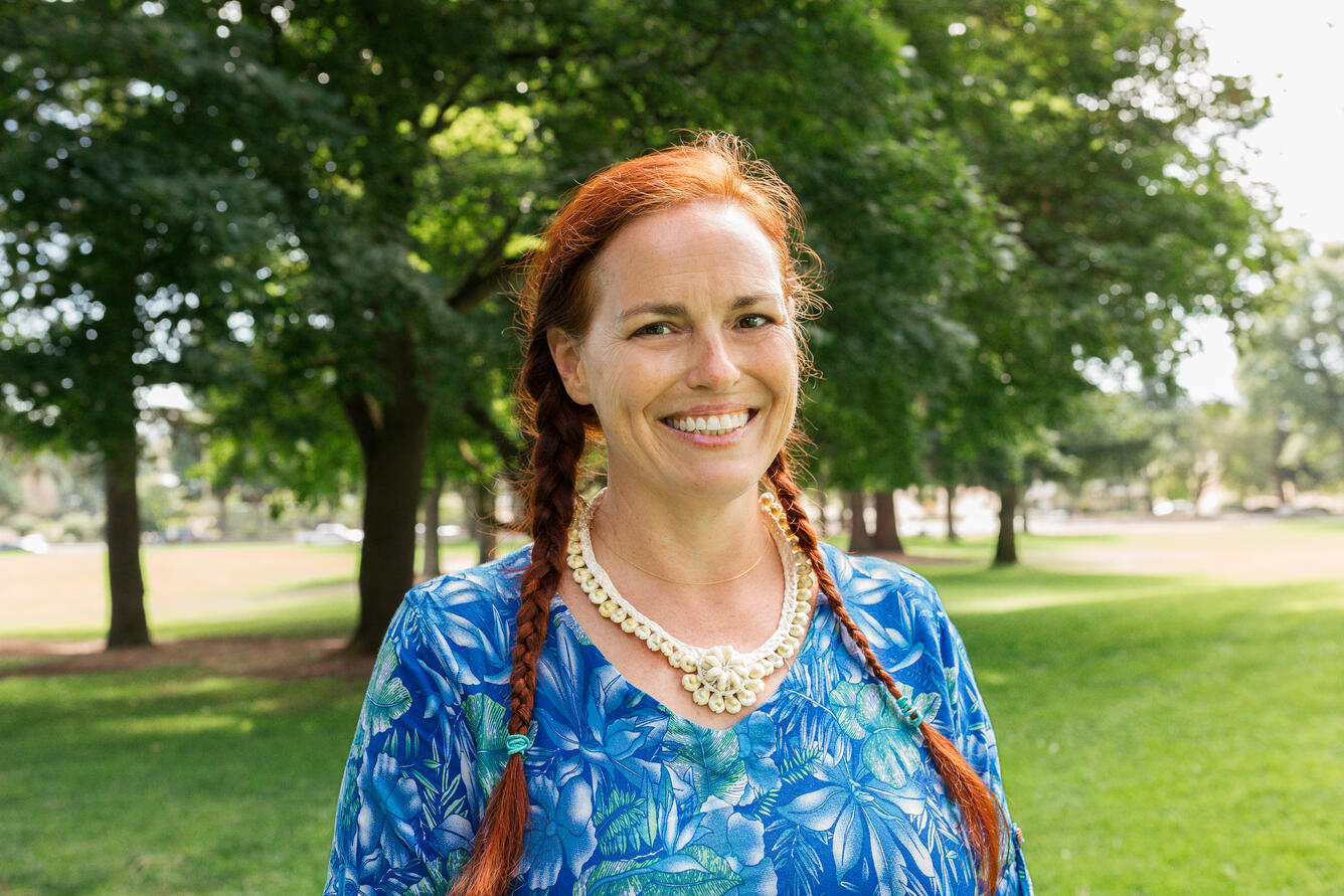 A woman with long dark, braided hair wearing a blue floral shirt smiles for her picture while in a park.