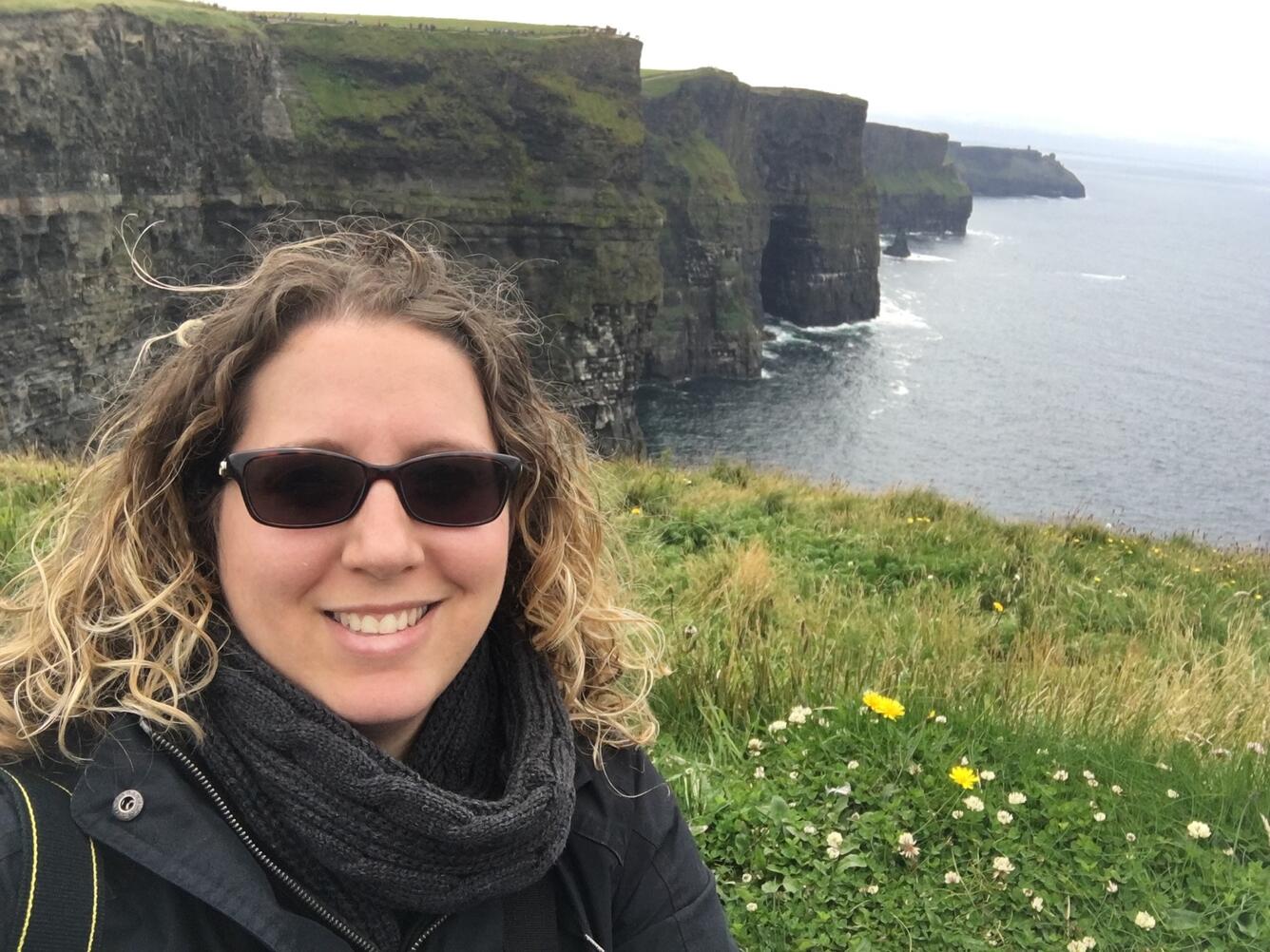 A person in sunglasses and a jacket and scarf smiles at the camera while standing along a rocky cliff covered in green grass and yellow and white flowers