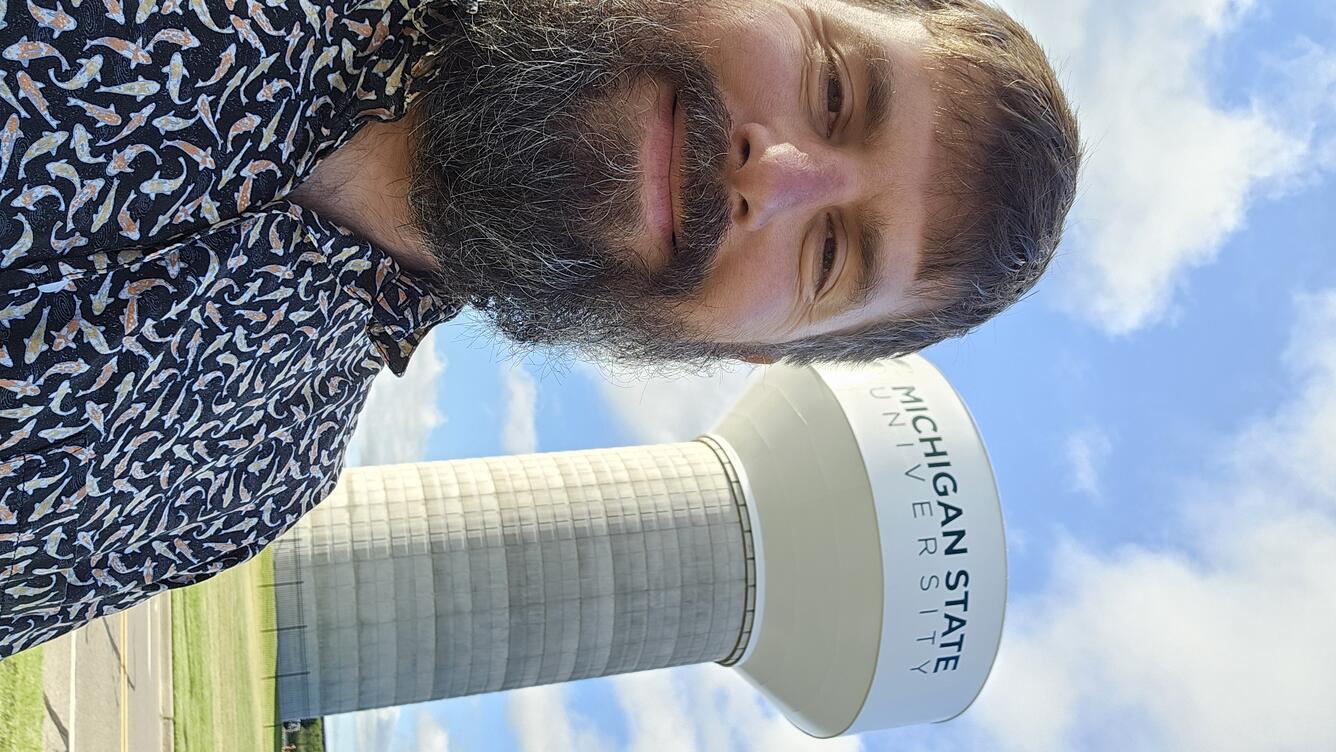 scientist wearing a fish print blue shirt with brown hair beard standing near a michigan state university water tower on a sunny day