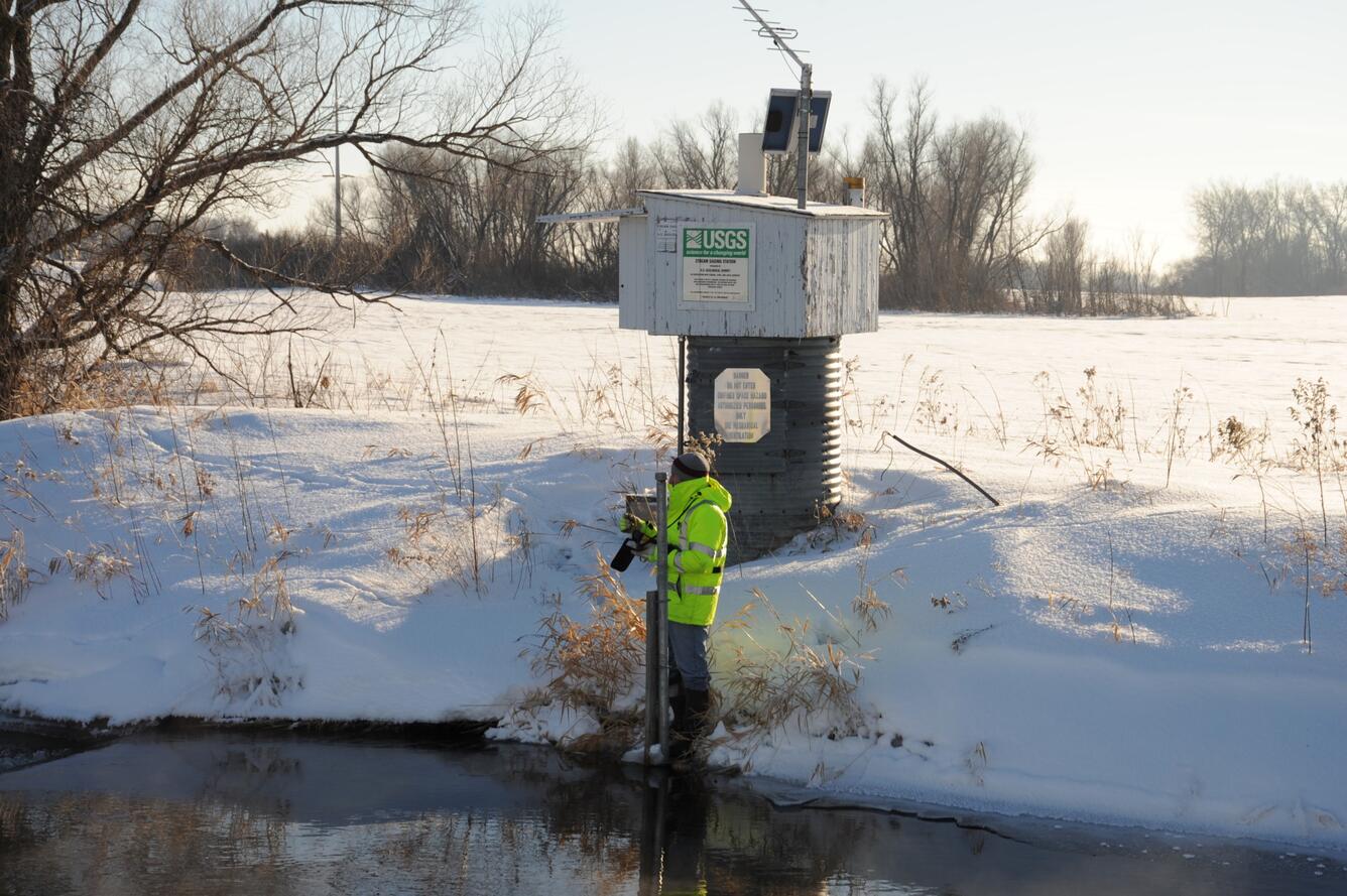 A hydrologic technician wearing a bright yellow coat stands in front of a streamgage housing and takes a streamflow reading