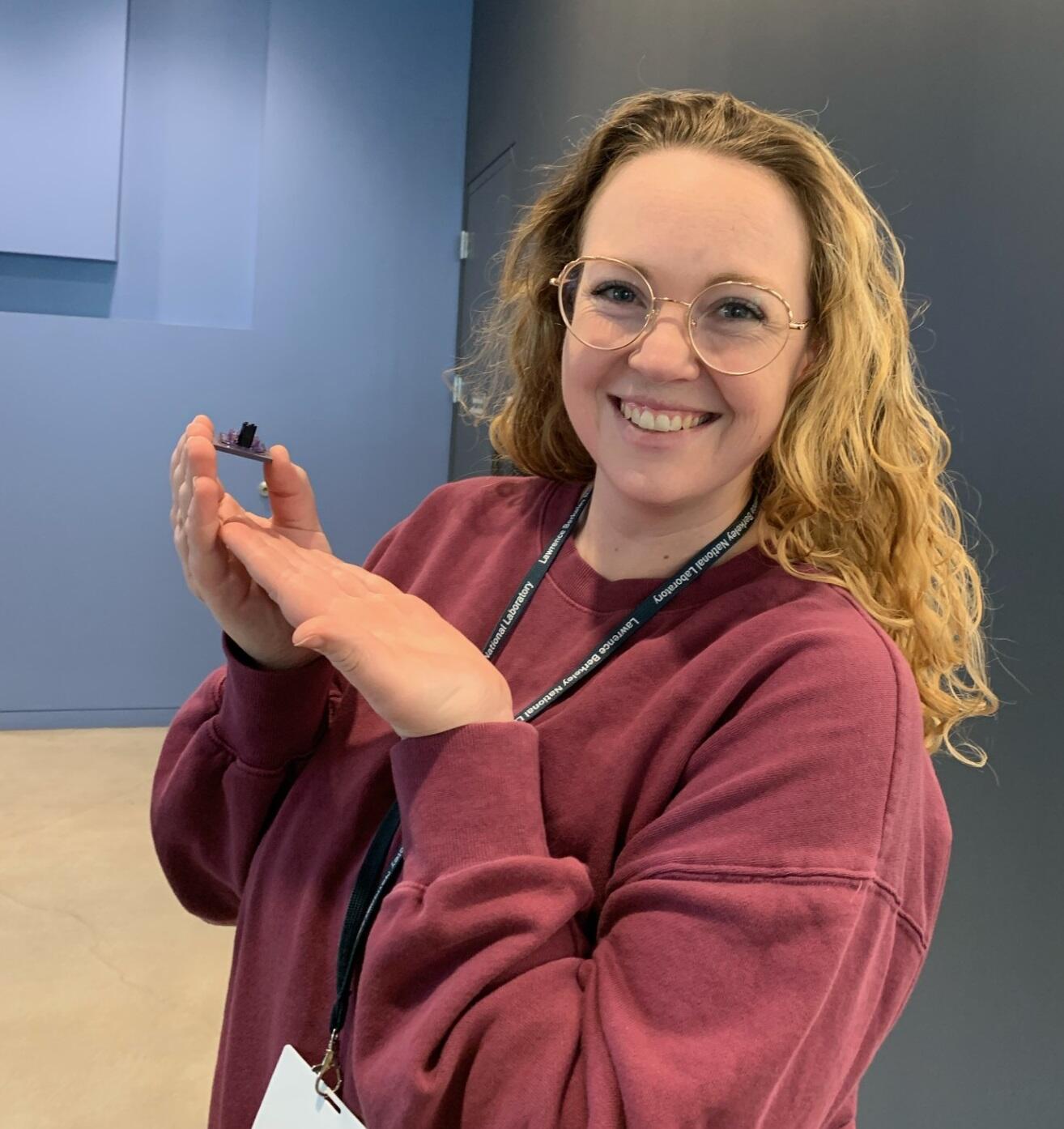 Woman with curly blonde hair wearing glasses smiling and holding small object