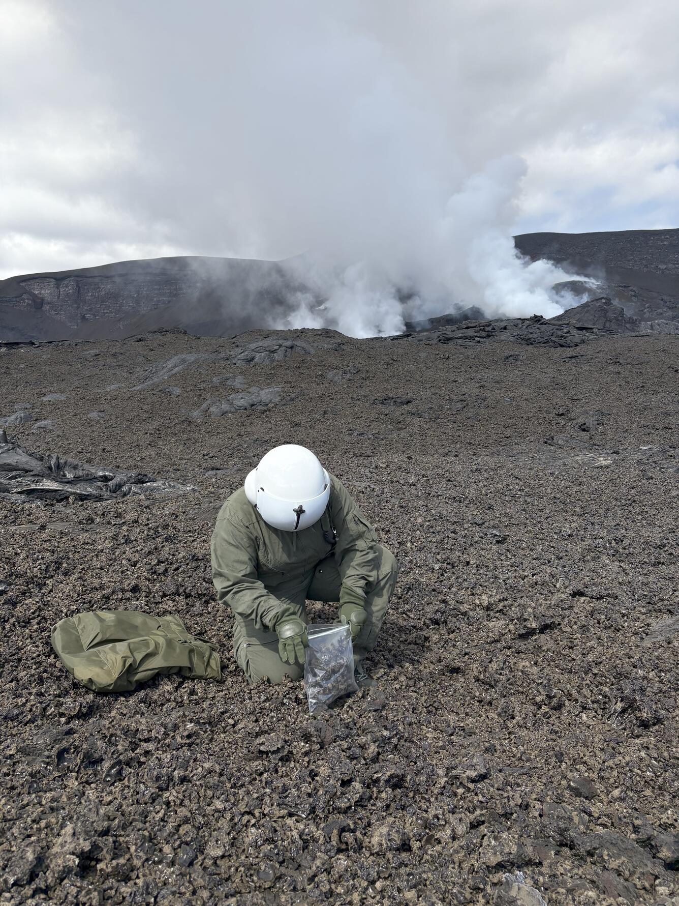 Color photograph of scientist collecting geologic samples near degassing volcanic vent