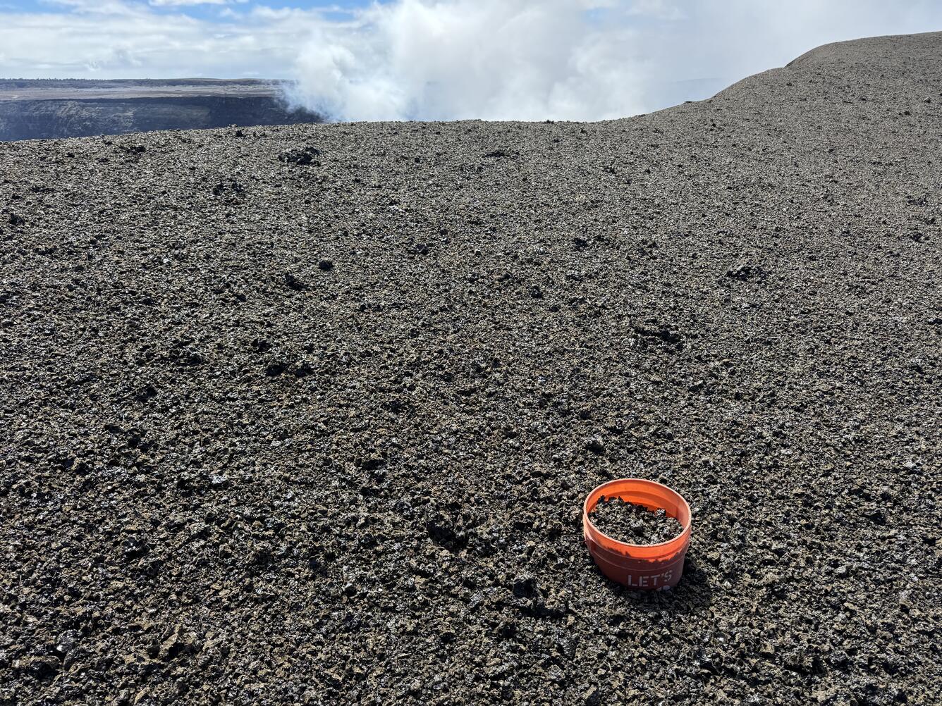 Color photograph of volcanic terrain with bucket in foreground and degassing in background