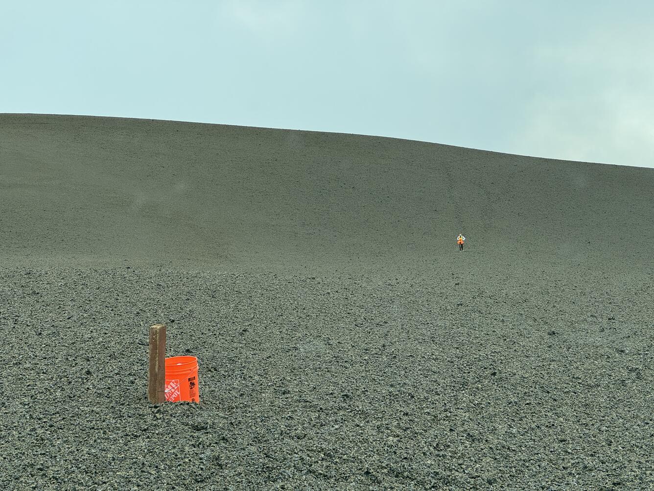 Color photograph of bucket next to mound of volcanic material with person walking in the distance