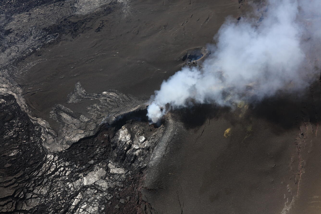 color photo looking down at cooled black lava and pale gray steam.