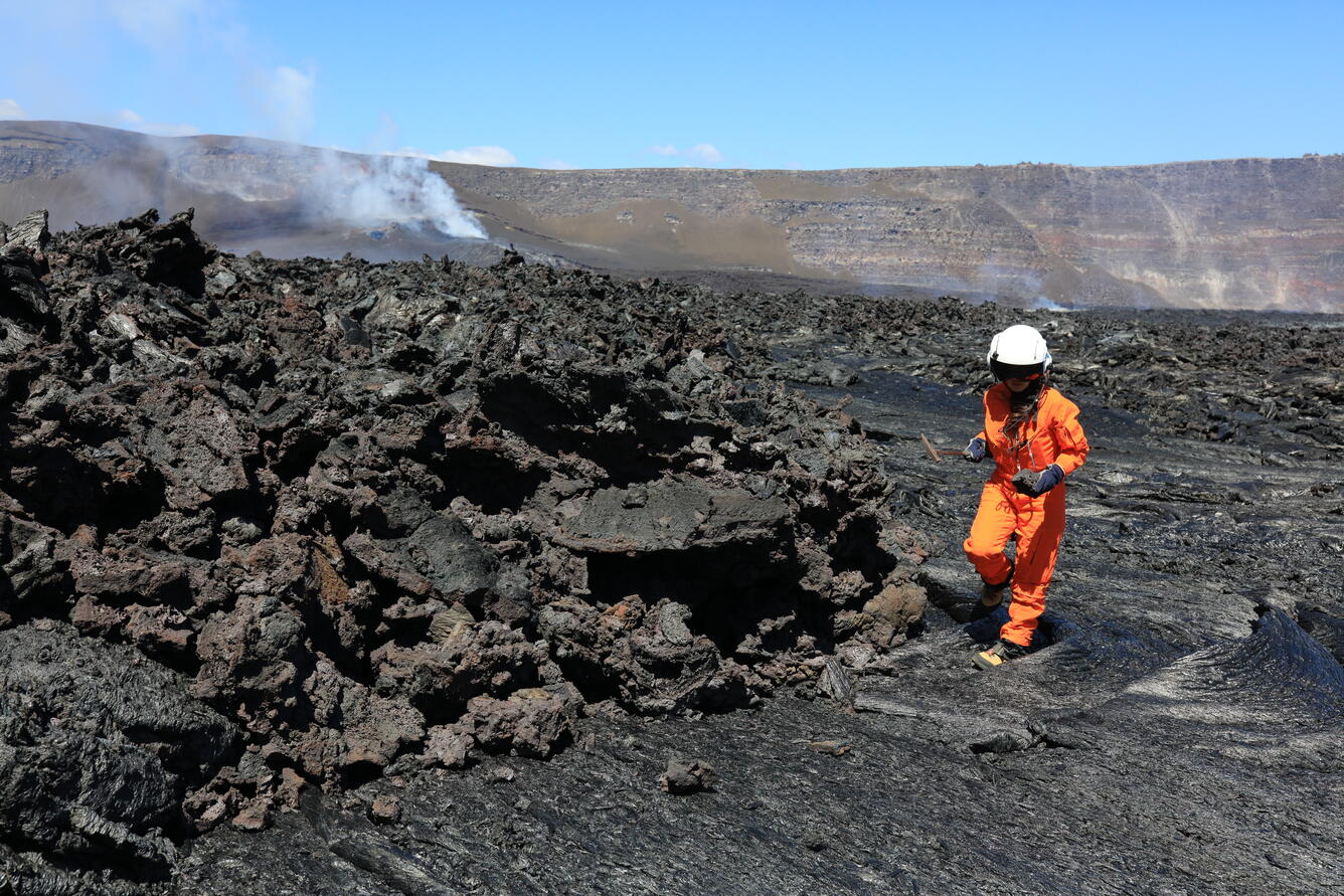 A scientists wearing and orange flight suit and a white flight helmet samples black, cooled lava. 