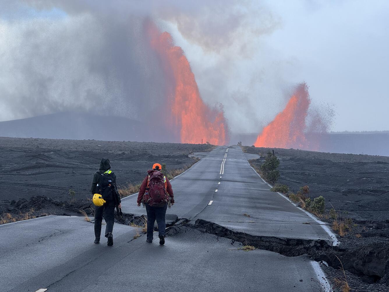 Color photograph of scientists walking on damaged road with lava fountains in the background