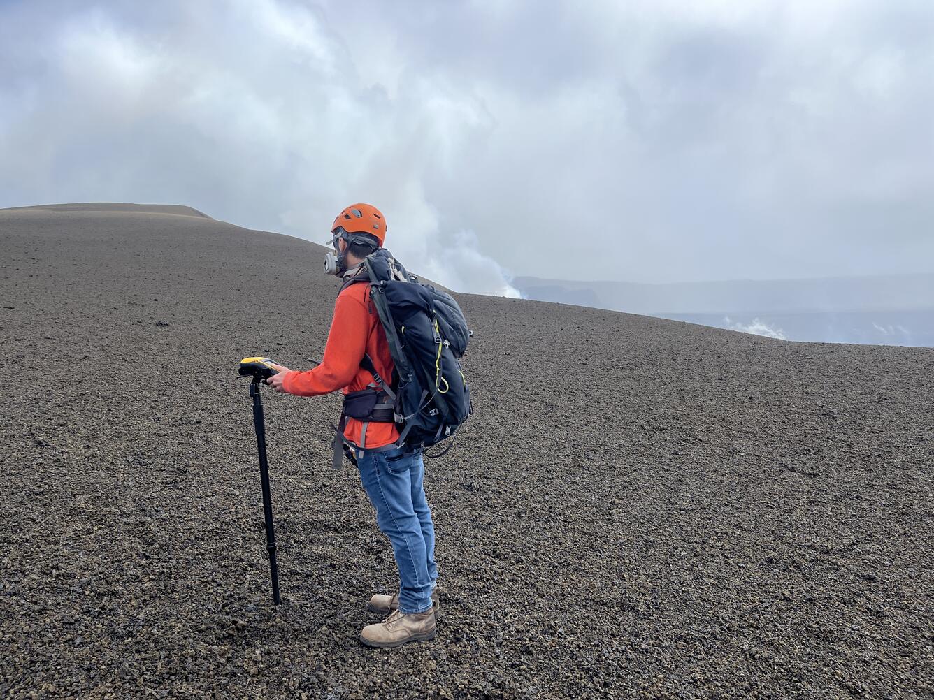 Color photograph of scientist wearing personal protective equipment and taking a measurement on a crater rim