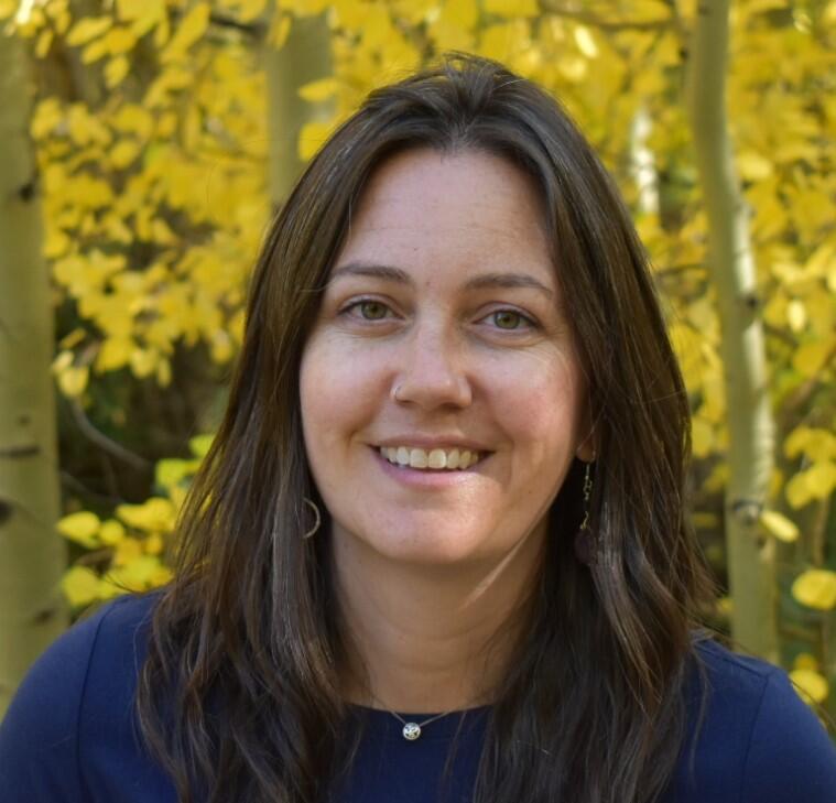 A woman with shoulder length brown hair smiles for her picture while standing in front of a thicket of autumn trees.