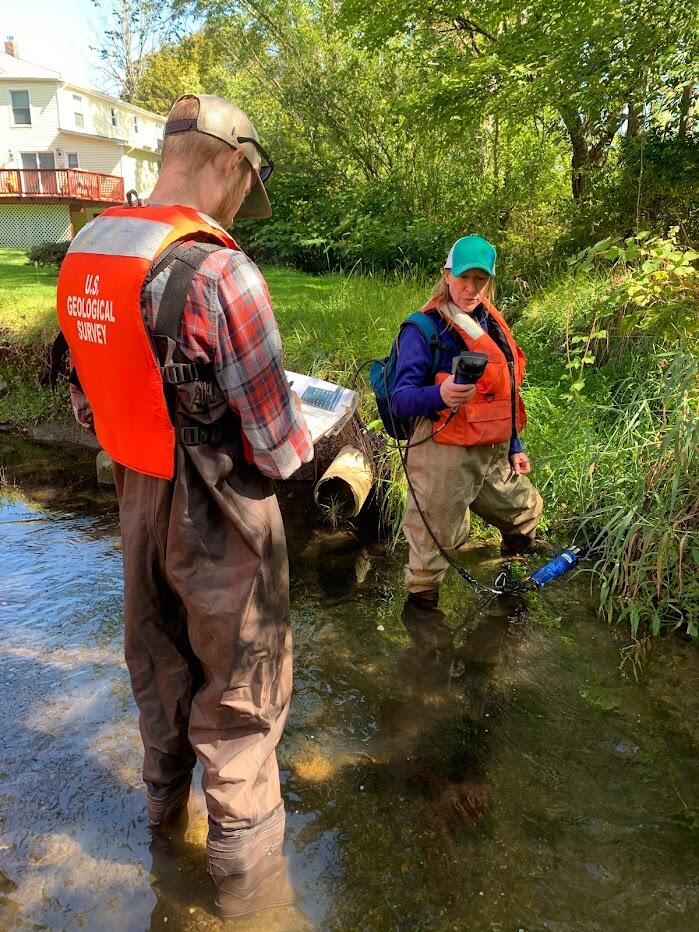 Two scientists in orange vests taking reading from a sampling probe in the edge of a river.