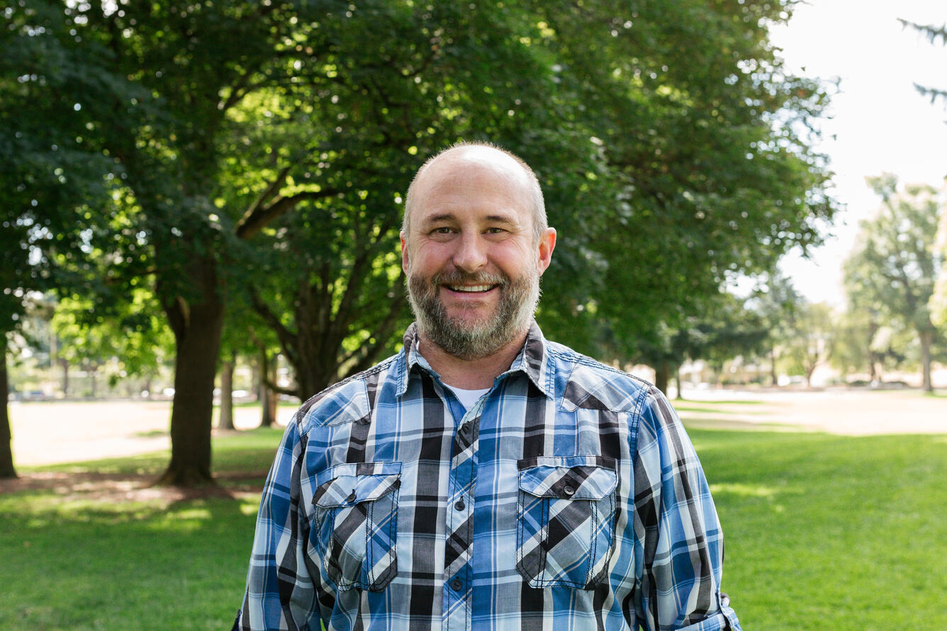 A man in a blue plaid button up smiles for his picture while standing in a park.