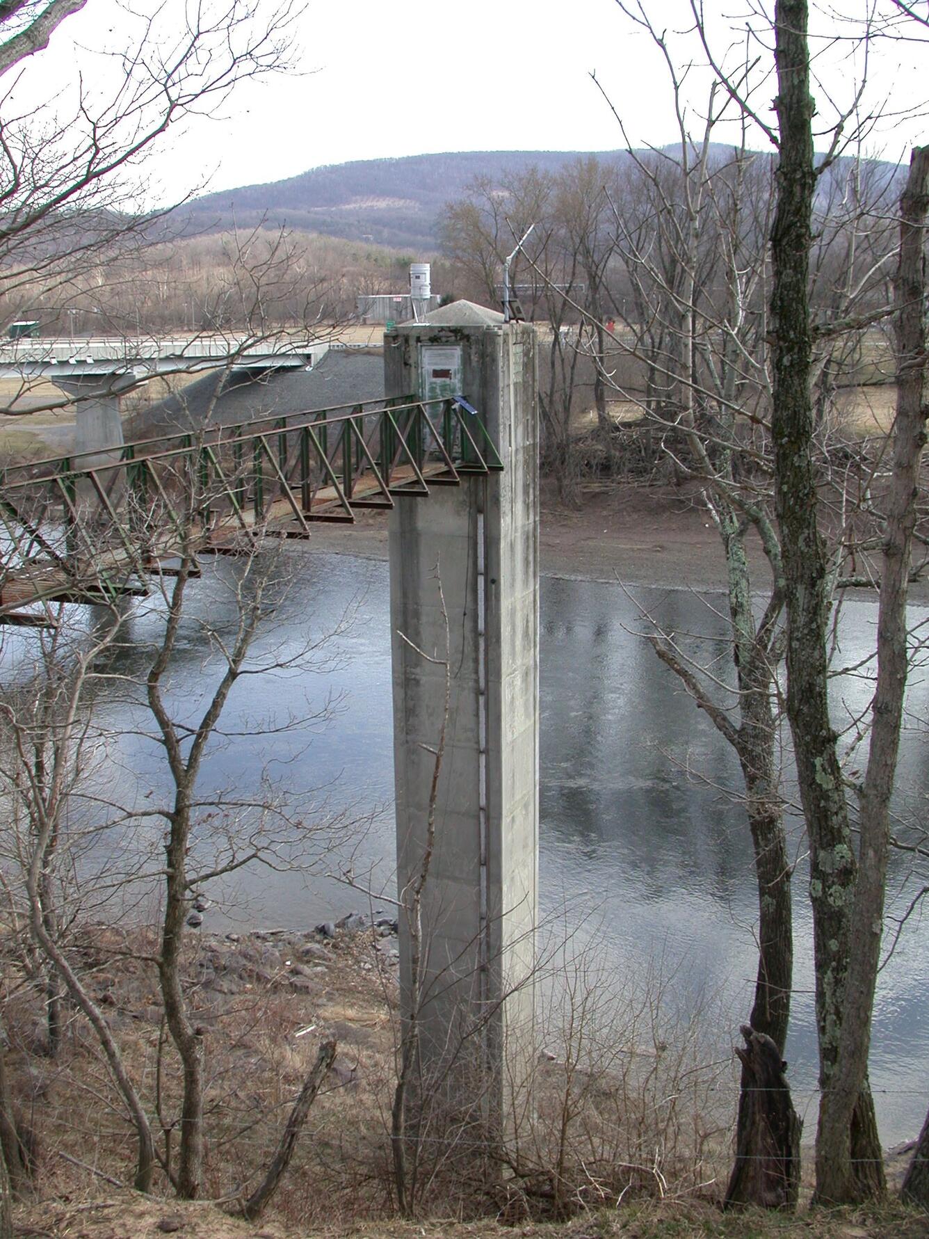 A bridge leads to a tall gage house adjacent to a river.