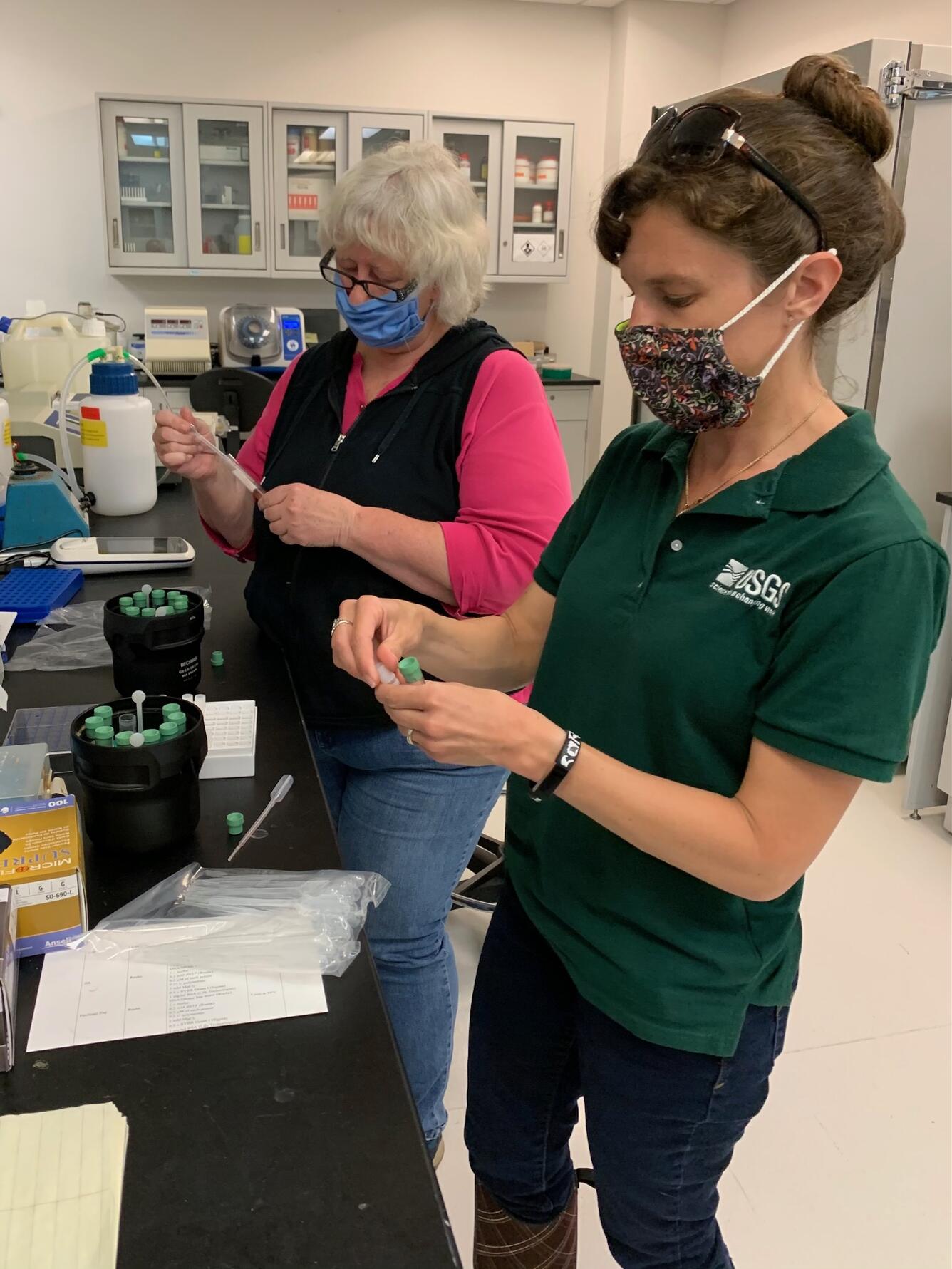 Two scientists wearing masks stand at a lab bench pipetting specimen tubes.