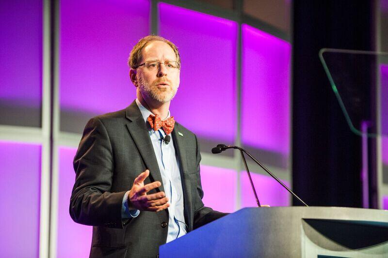 Image shows a man in a bowtie speaking at a podium