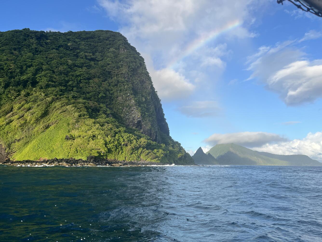 Color photo of a large green cliff on an island towering over the blue ocean. 