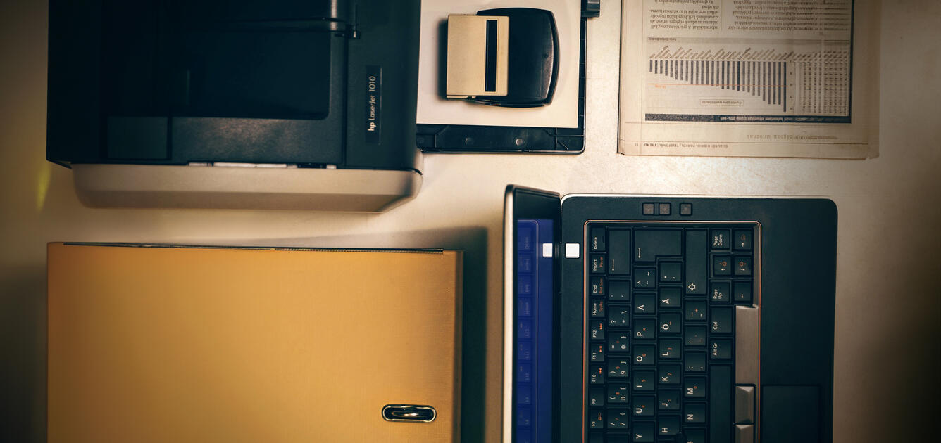 Image of desktop with printer, stamp, newspaper, and open laptop