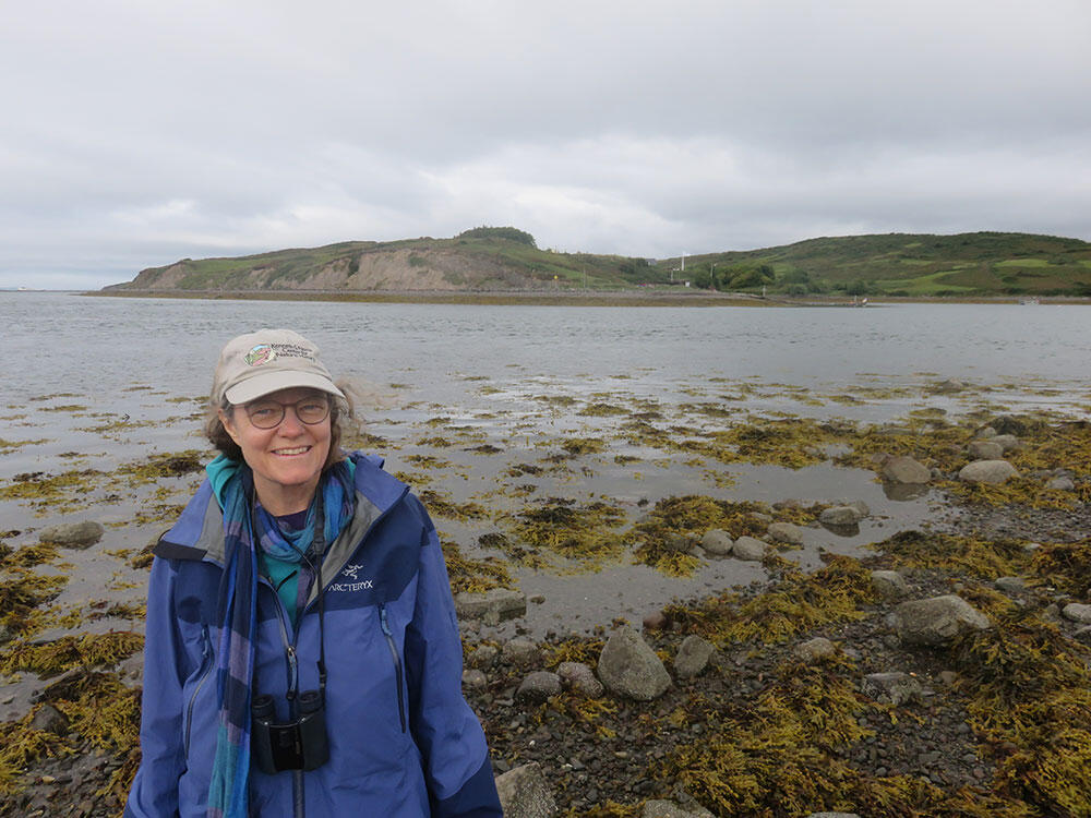 A woman wearing a hat and jacket and eyeglasses smiles for the camera at a rocky beach with cliffs in background.