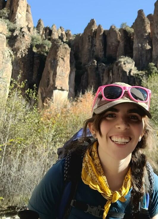 Woman wearing a hat and bandana smiles at camera, with red rock formations in the background