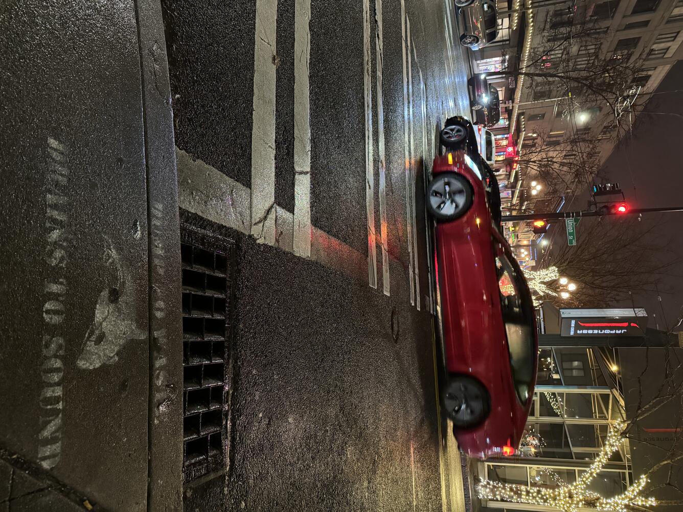 Seattle storm drain in the rain with a sign that reads "Dump no waste, drains to sound" and a red car passing by