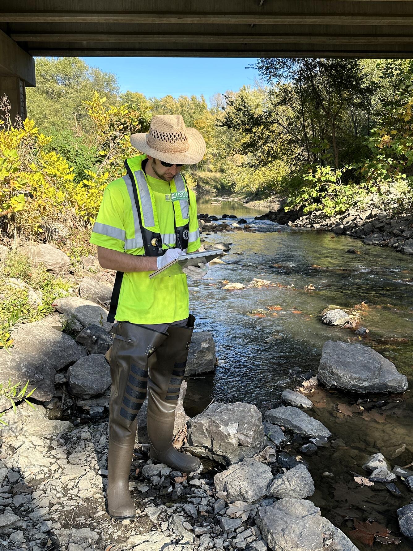 Scientist records conditions of river while standing next to it with a highway overpass in background