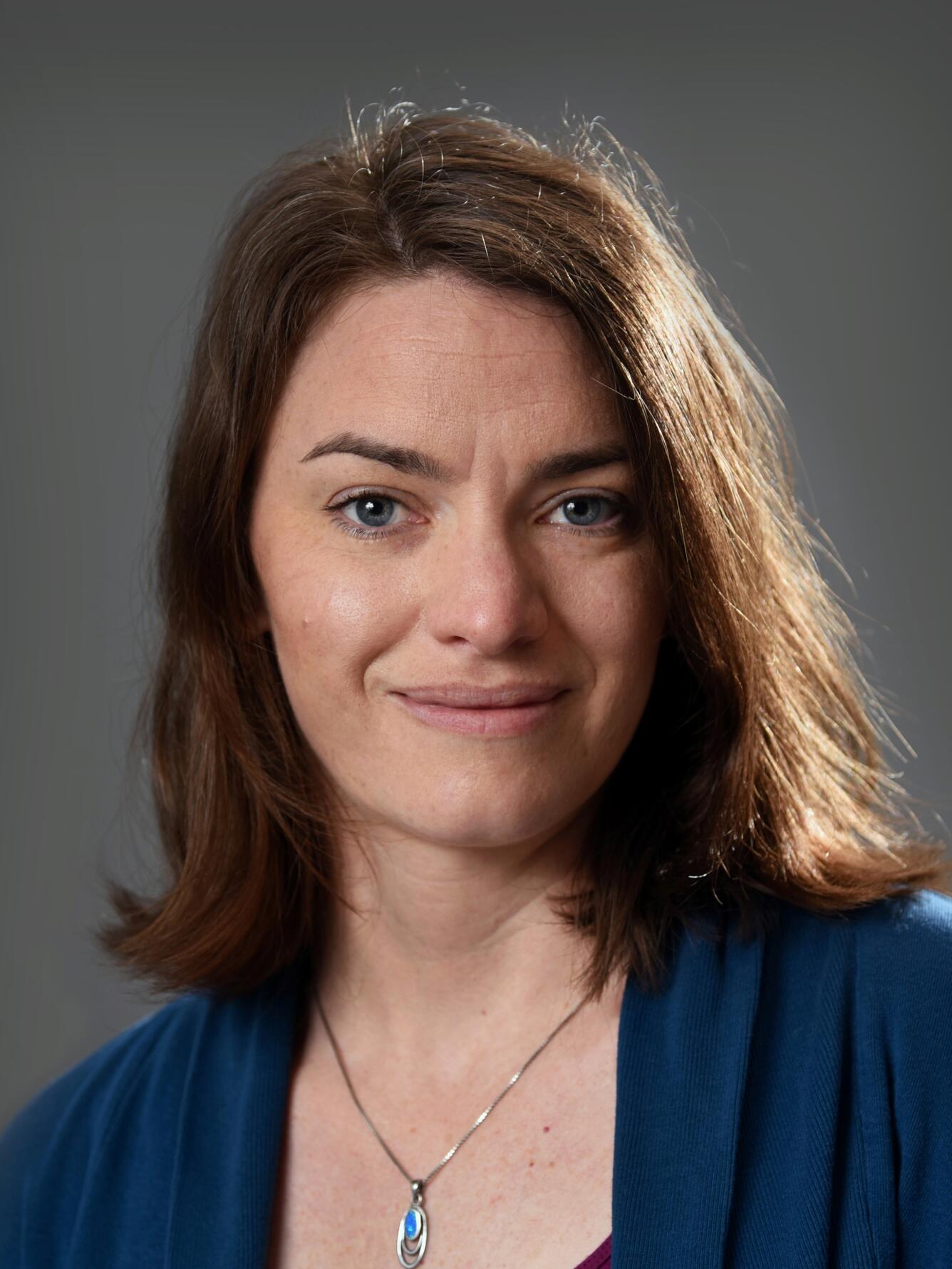 scientist with shoulder-length brown hair, blue eyes, wearing a blue necklace and wearing a teal sweater
