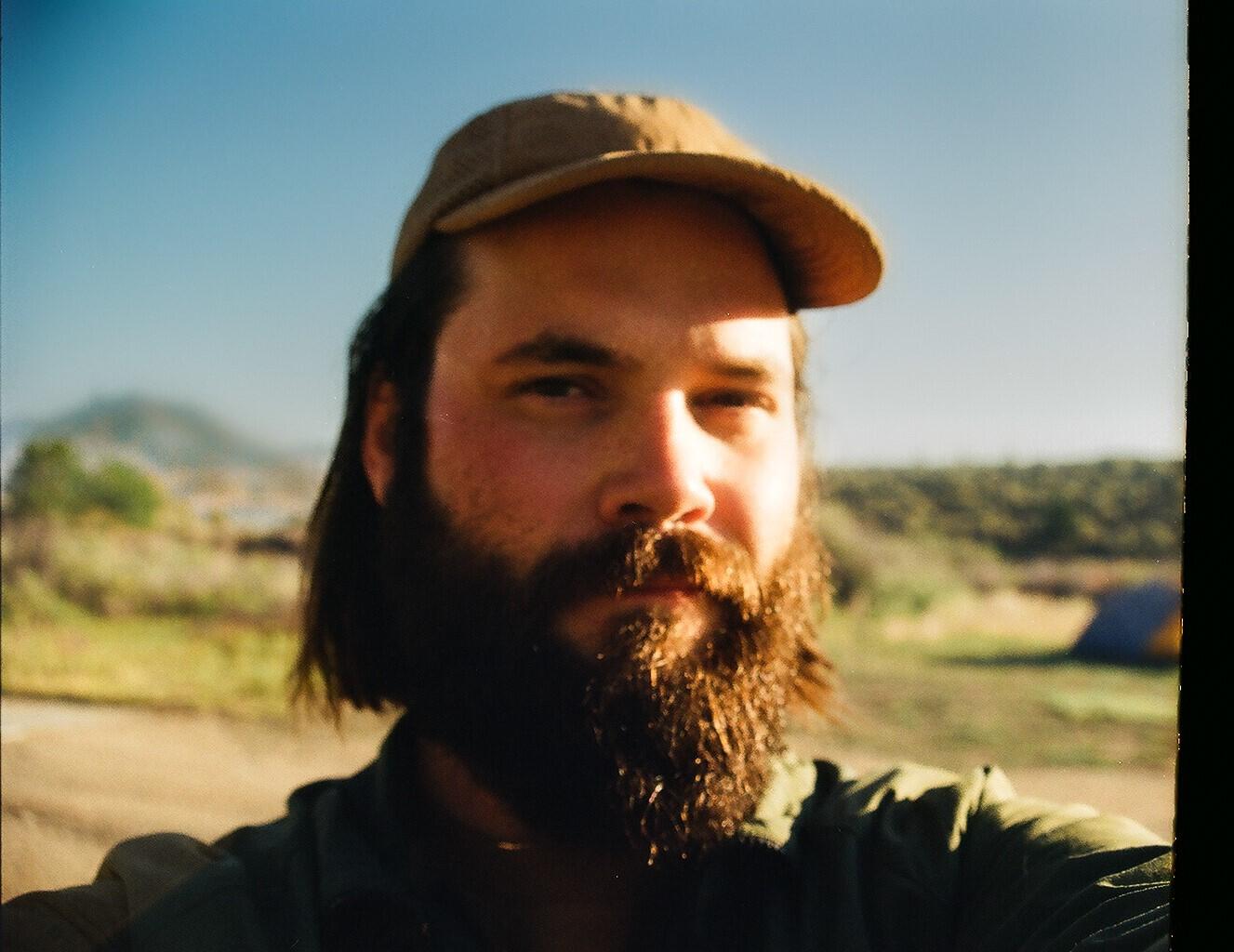 headshot of a male with dark brown beard and tan hat. Outdoors on a sunny day.