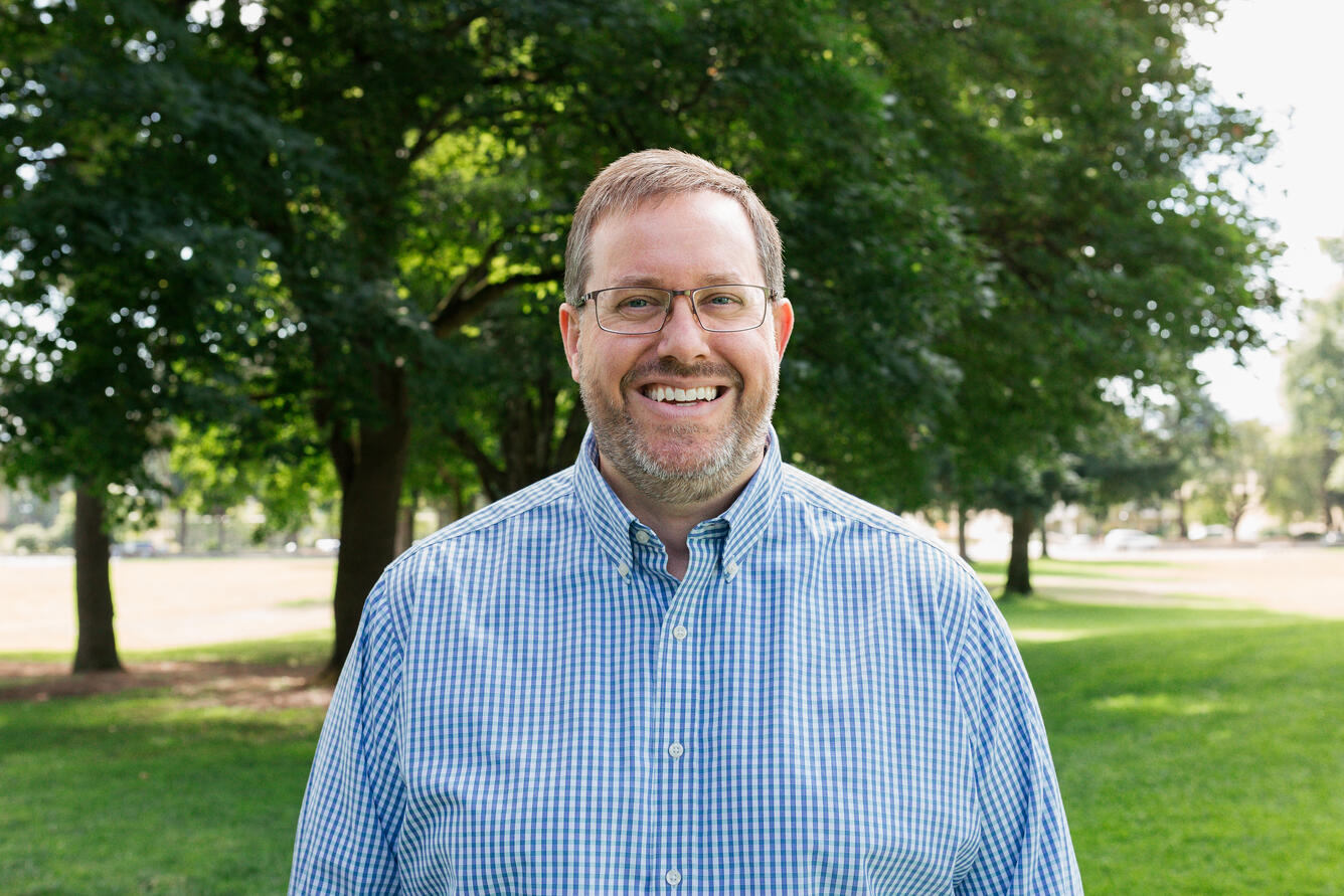 A man with short hair and glasses wearing a blue button up shirt smiles for his picture while standing in a park.