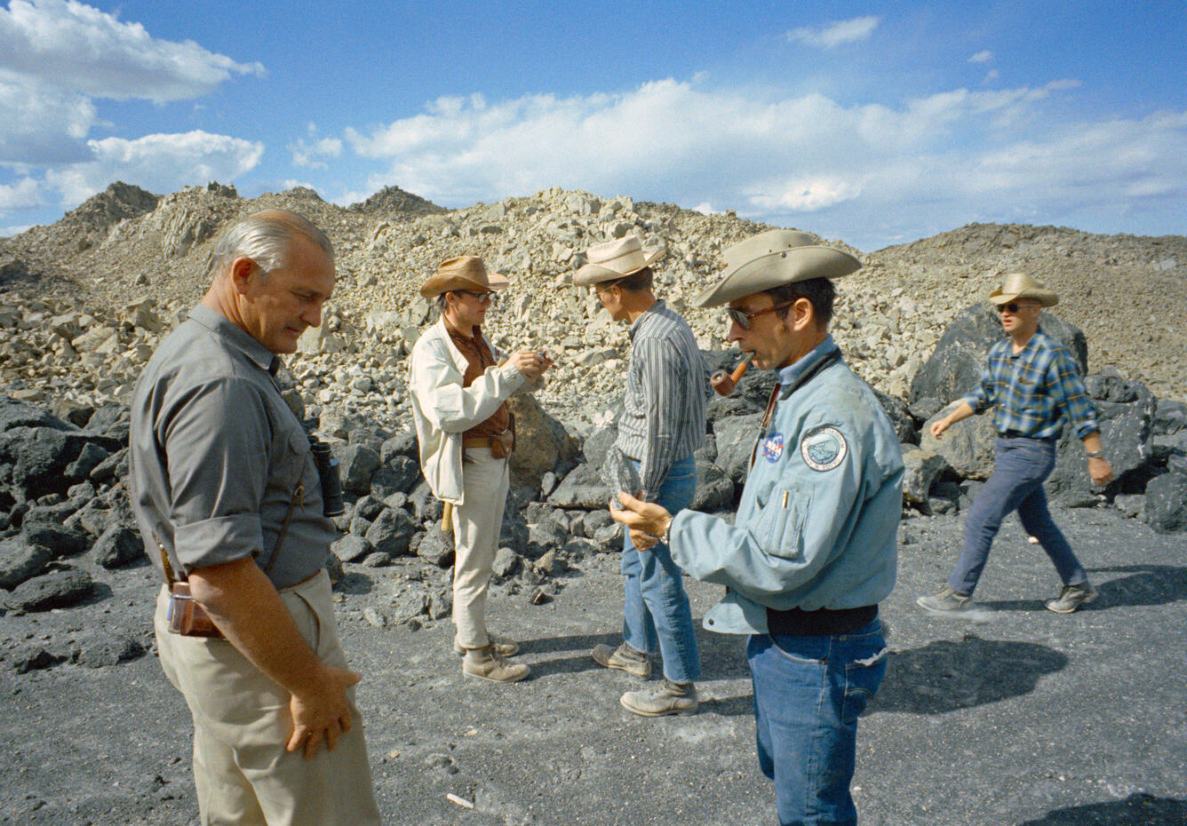 A group of five white men wearing cowboy hats, jeans or khakis, and button-down shirts are standing on a sandy gray surface ringed by large gray boulders of volcanic rock. In the background, a number of bare, rubbly, rocky peaks are lit by afternoon sun. The men are examining hand samples of rocks and one is smoking a pipe.