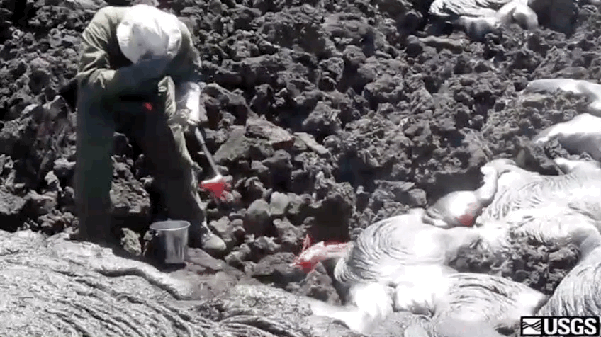 A person scoops lava from a flow with a rock hammer while wearing protective gear and shielding their face.