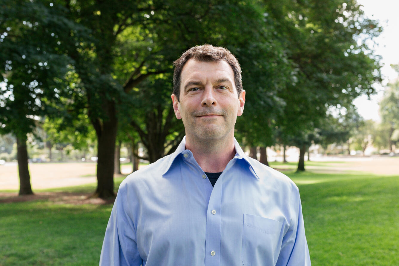 A man with short brown hair and a blue button up shirt standing in park. 