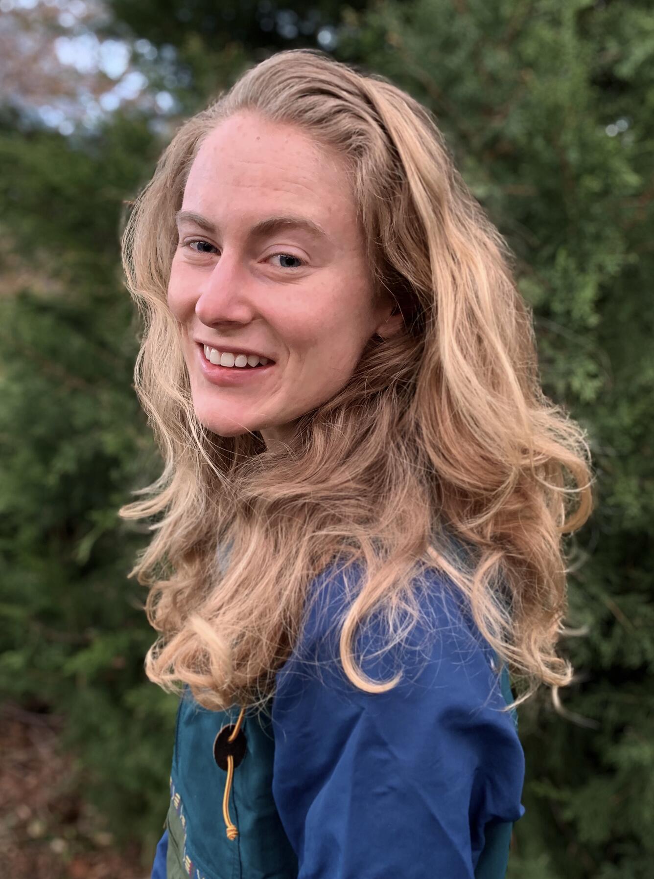 Photo of blonde woman in blue shirt in front of greenery