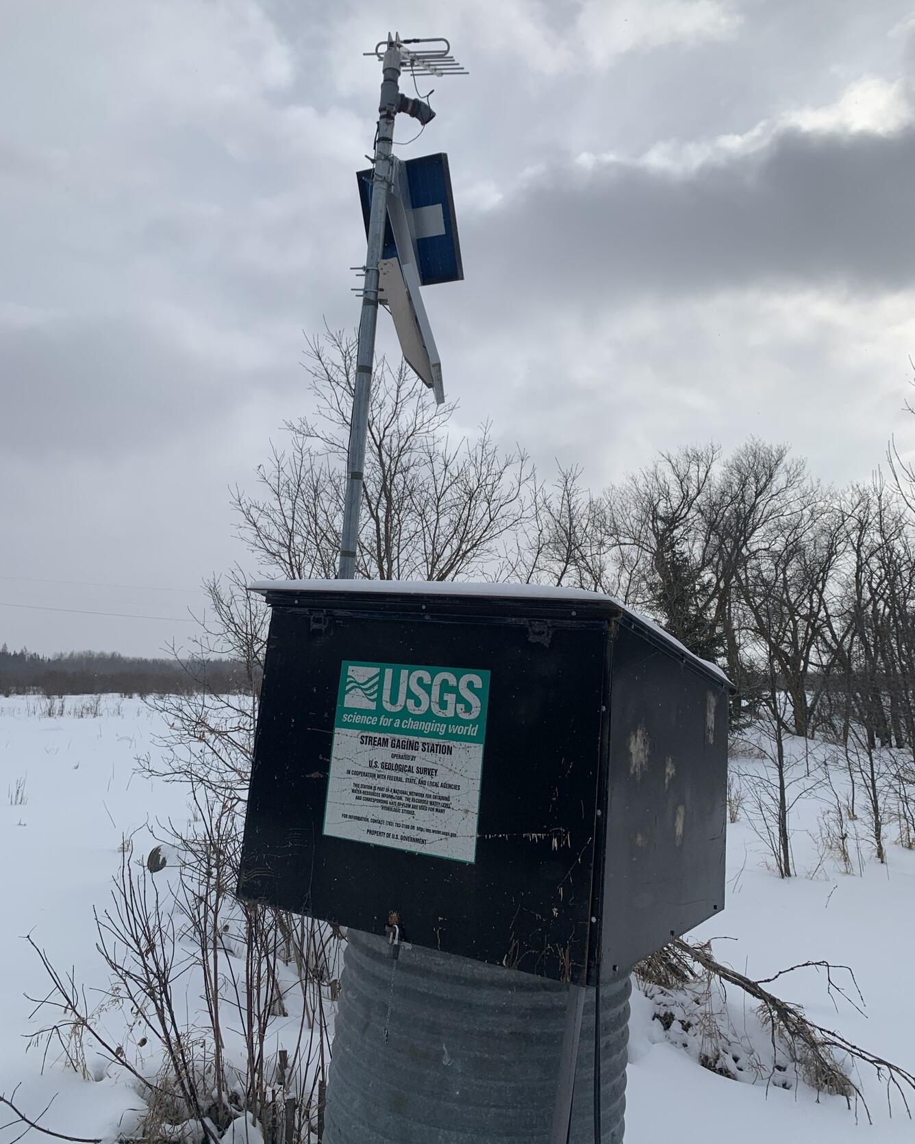USGS streamgage housing with solar panel and sensors mounted on a pole, against a snow-covered rural background