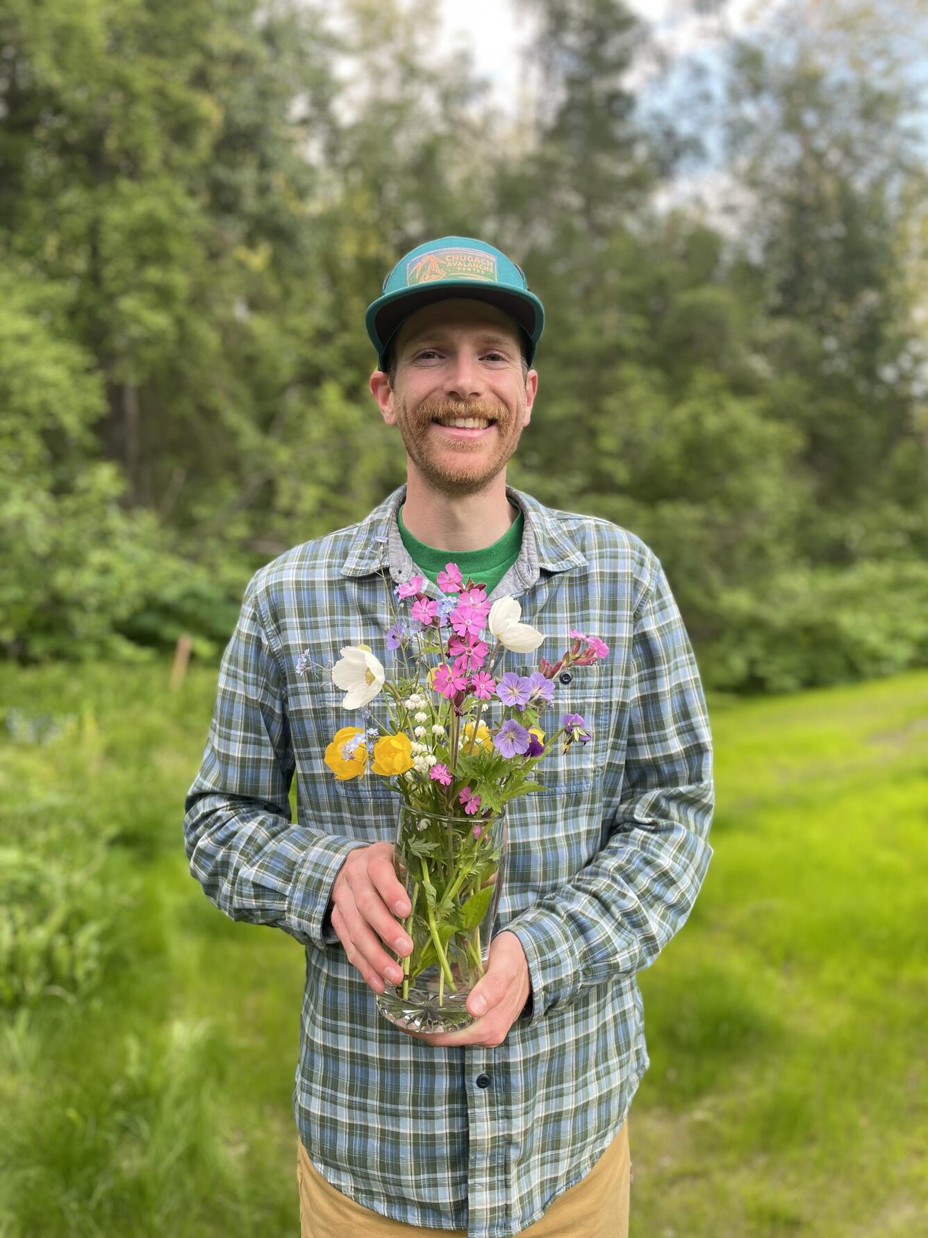 A man wearing a plaid button up and a baseball cap holds a bouquet of flowers.