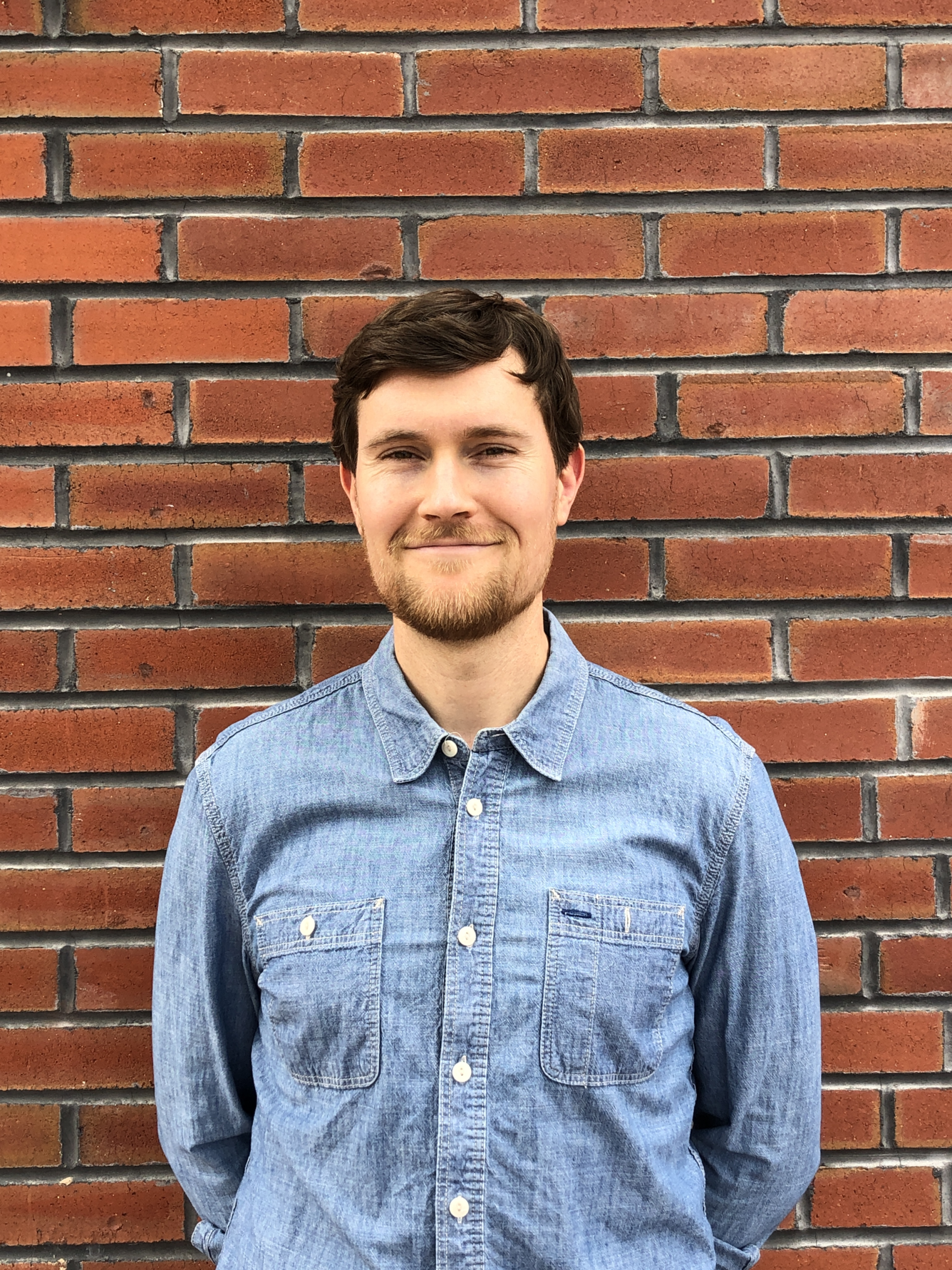 A man in a blue button up shirt with short brown hair stands in front of a brick wall for his picture. 