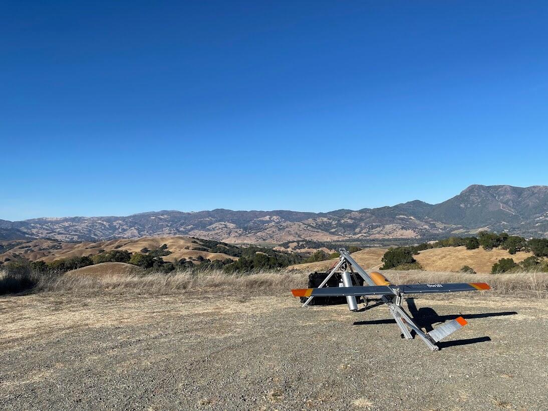 A large grey and orange drone mounted on a launch pad stationed in an open field with rolling hills in the background.