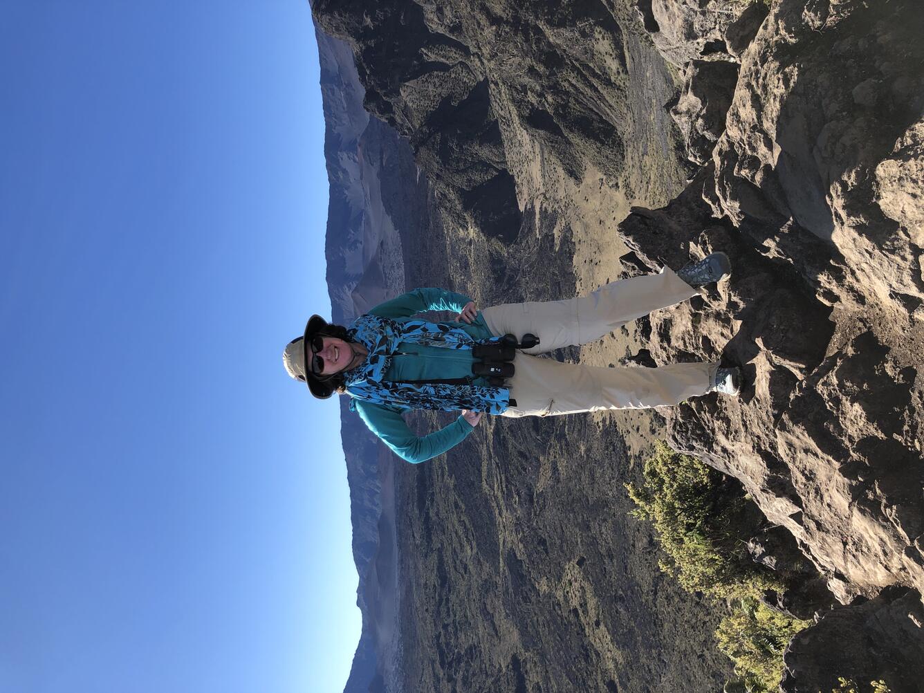 researcher wearing a green shirt, tan pants and hat, black sunglasses, standing on a large rock